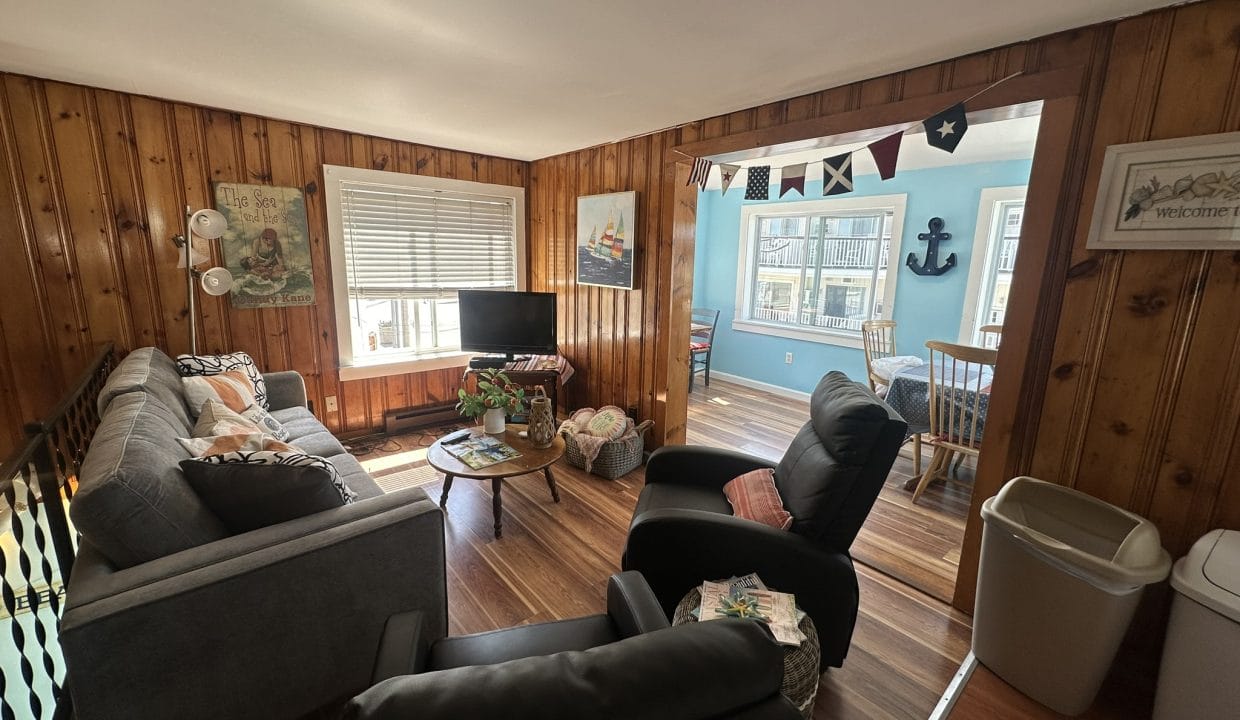 A cozy living room with wood-paneled walls features a sofa, armchairs, a small TV, and a coffee table. An adjacent dining area with nautical decor is visible through a wide doorway.