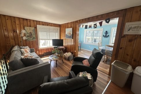 A cozy living room with wood-paneled walls features a sofa, armchairs, a small TV, and a coffee table. An adjacent dining area with nautical decor is visible through a wide doorway.