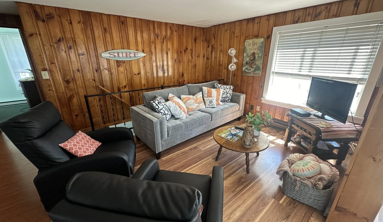 A cozy living room with wooden walls, gray sofa, two black chairs, a round coffee table, and a TV. Surf sign and decorative pillows are visible.