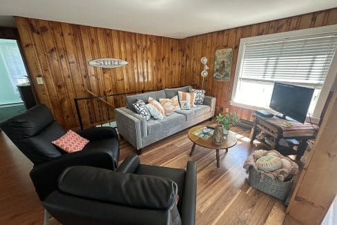 A cozy living room with wooden walls, gray sofa, two black chairs, a round coffee table, and a TV. Surf sign and decorative pillows are visible.