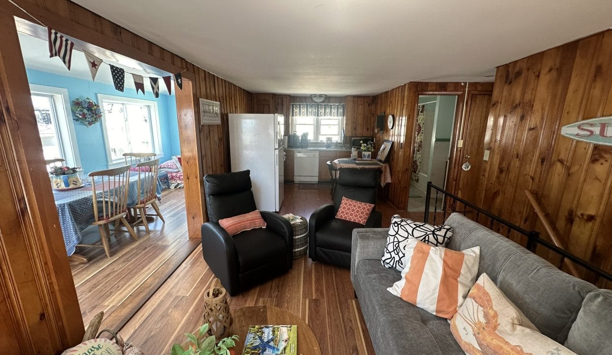 A cozy living room with wooden walls, a gray sofa, two black recliners, and a view into a kitchen and dining area with colorful decor. A table with magazines and plants is in the foreground.