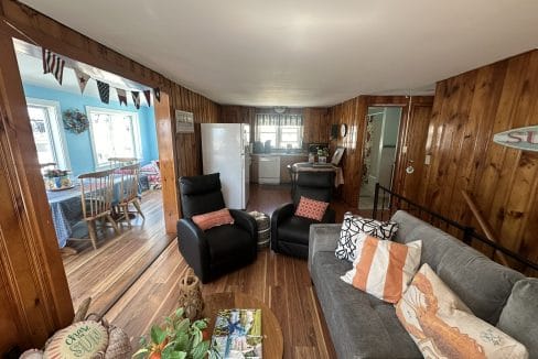 A cozy living room with wooden walls, a gray sofa, two black recliners, and a view into a kitchen and dining area with colorful decor. A table with magazines and plants is in the foreground.