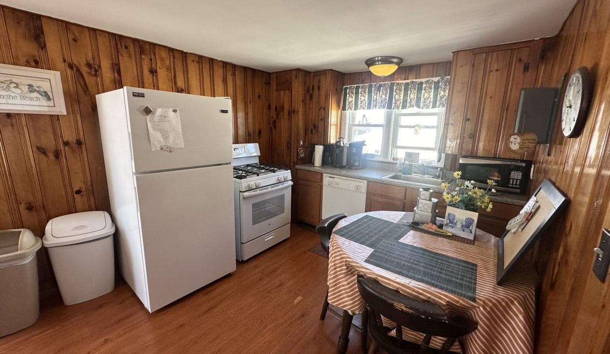 A kitchen with wood-paneled walls, a white refrigerator, stove, dishwasher, and a table with a checkered cloth. Two chairs surround the table, and various appliances are on the counter.