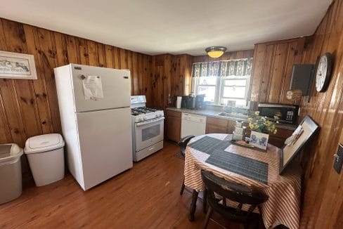 A kitchen with wood-paneled walls, a white refrigerator, stove, dishwasher, and a table with a checkered cloth. Two chairs surround the table, and various appliances are on the counter.