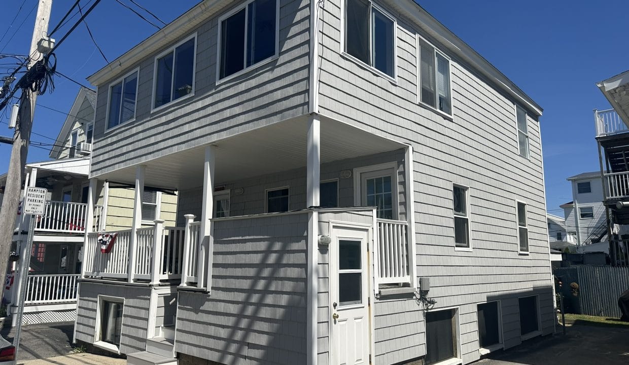 Two-story gray house with white trim, featuring a covered front porch and a side entrance, set under a clear blue sky.