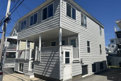 Two-story gray house with white trim, featuring a covered front porch and a side entrance, set under a clear blue sky.