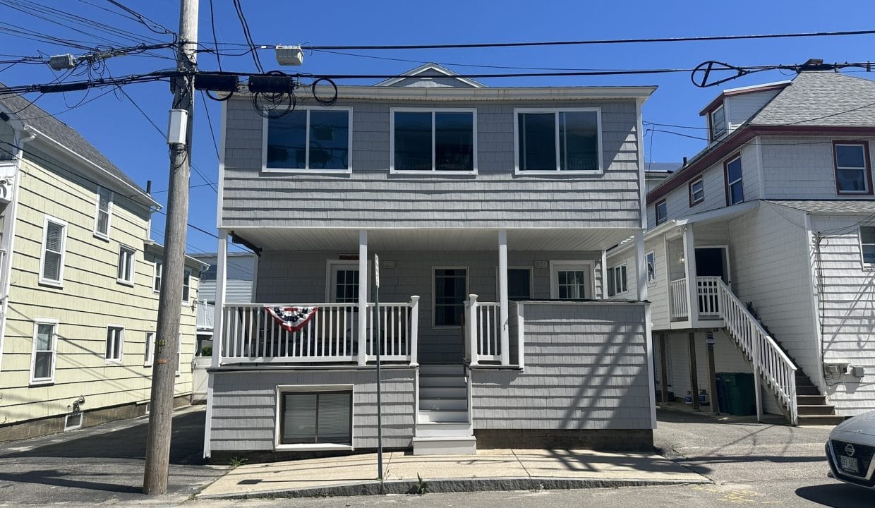 A two-story gray house with a small porch, large windows, and a patriotic banner. It is located on a street with overhead wires and neighboring houses on either side.