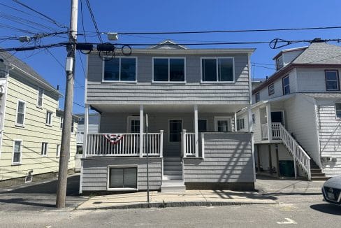 A two-story gray house with a small porch, large windows, and a patriotic banner. It is located on a street with overhead wires and neighboring houses on either side.