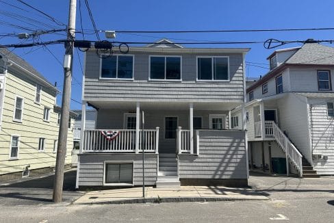 Two-story gray house with a front porch and driveway, flanked by power lines and neighboring houses, under a clear blue sky.