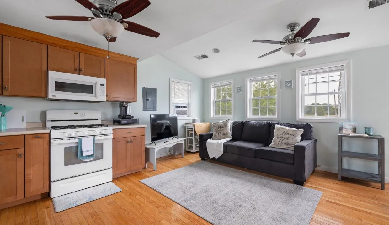 Small living space with a kitchen area. Features wooden cabinets, a stove, microwave, a black sofa, two ceiling fans, and a TV. Three windows provide natural light.