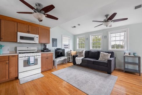 Small living space with a kitchen area. Features wooden cabinets, a stove, microwave, a black sofa, two ceiling fans, and a TV. Three windows provide natural light.