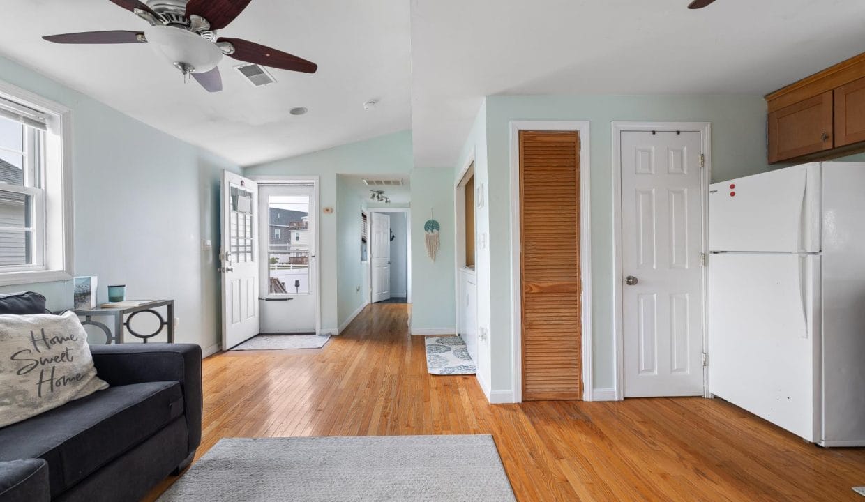 A living room with wooden floors, a black couch, a ceiling fan, and a white fridge next to a wooden door. An open front door leads to a hallway.