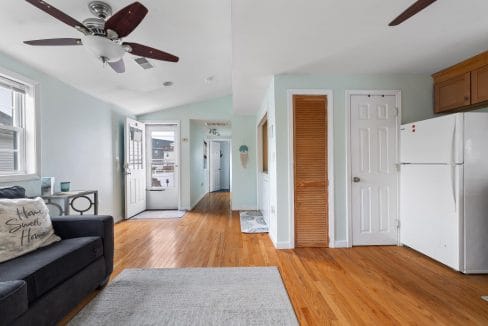 A living room with wooden floors, a black couch, a ceiling fan, and a white fridge next to a wooden door. An open front door leads to a hallway.