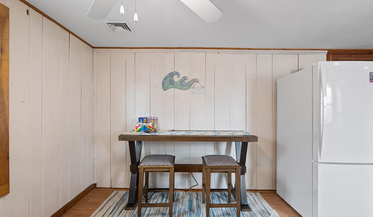 A small dining area with a wooden table, two stools, and a white fridge. A wave-themed artwork is on the wall above the table. A ceiling fan is visible at the top.