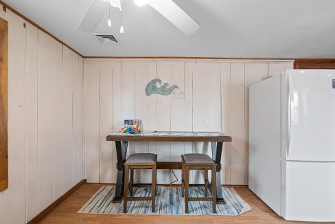 A small dining area with a wooden table, two stools, and a white fridge. A wave-themed artwork is on the wall above the table. A ceiling fan is visible at the top.