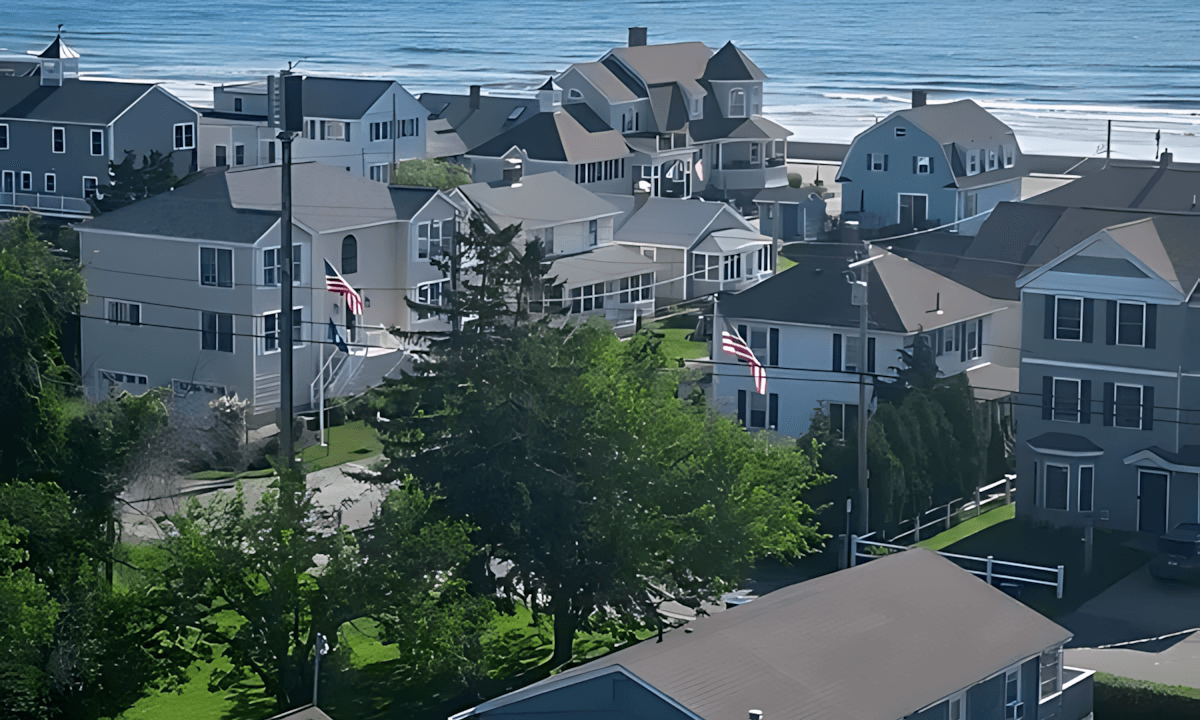 A coastal neighborhood with several houses near the ocean on a clear day. The scene includes greenery and distant ships on the horizon.