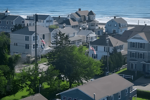 A coastal neighborhood with several houses near the ocean on a clear day. The scene includes greenery and distant ships on the horizon.