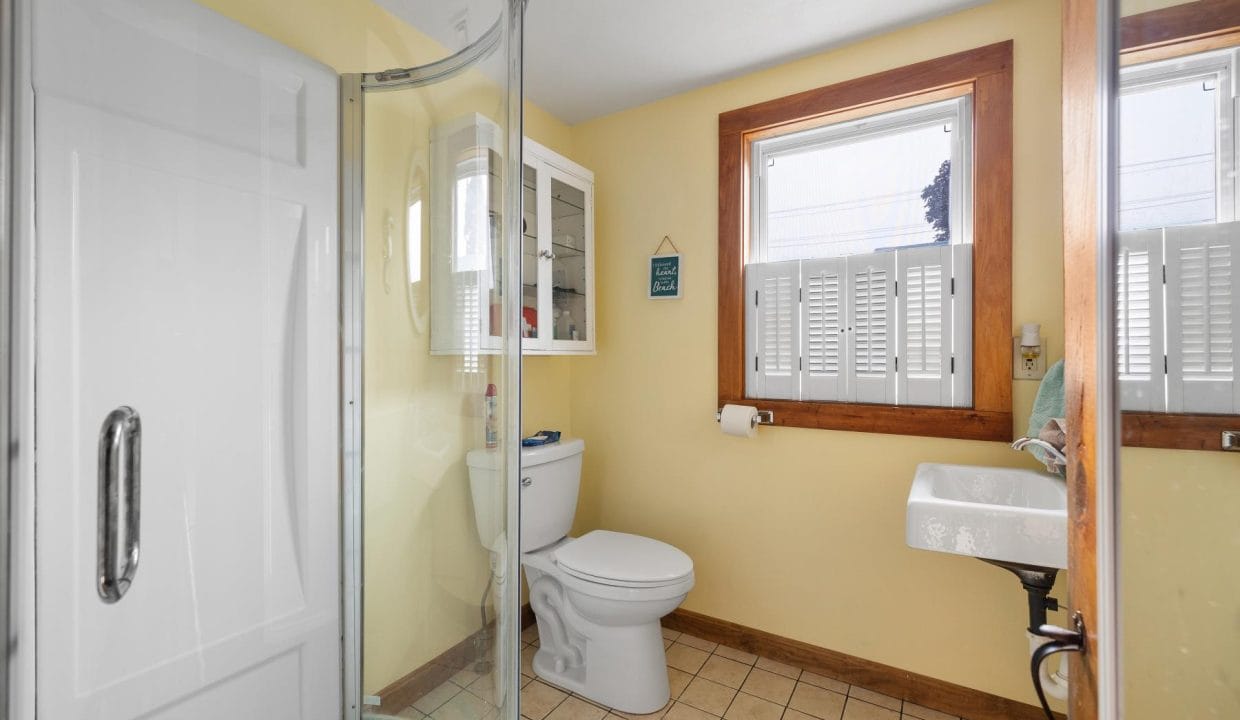 Small bathroom with a yellow-painted wall, corner shower, toilet, sink, and two windows with shutters. A cabinet is mounted above the toilet.