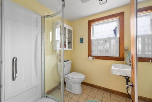 Small bathroom with a yellow-painted wall, corner shower, toilet, sink, and two windows with shutters. A cabinet is mounted above the toilet.