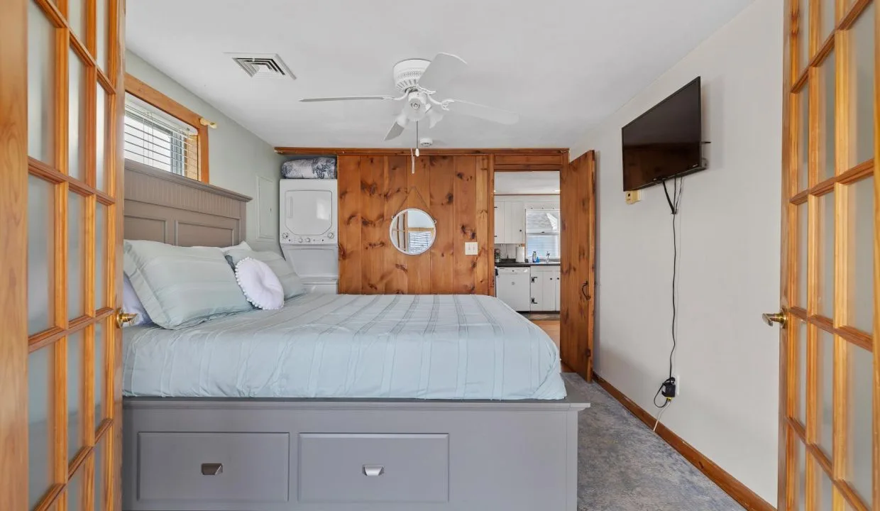 Bedroom with a large bed, ceiling fan, wall-mounted TV, mirrored closet, and laundry appliances in wooden cabinetry.