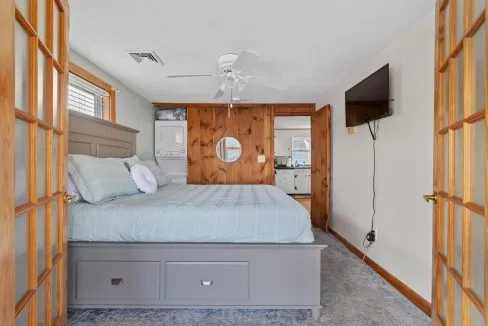 Bedroom with a large bed, ceiling fan, wall-mounted TV, mirrored closet, and laundry appliances in wooden cabinetry.