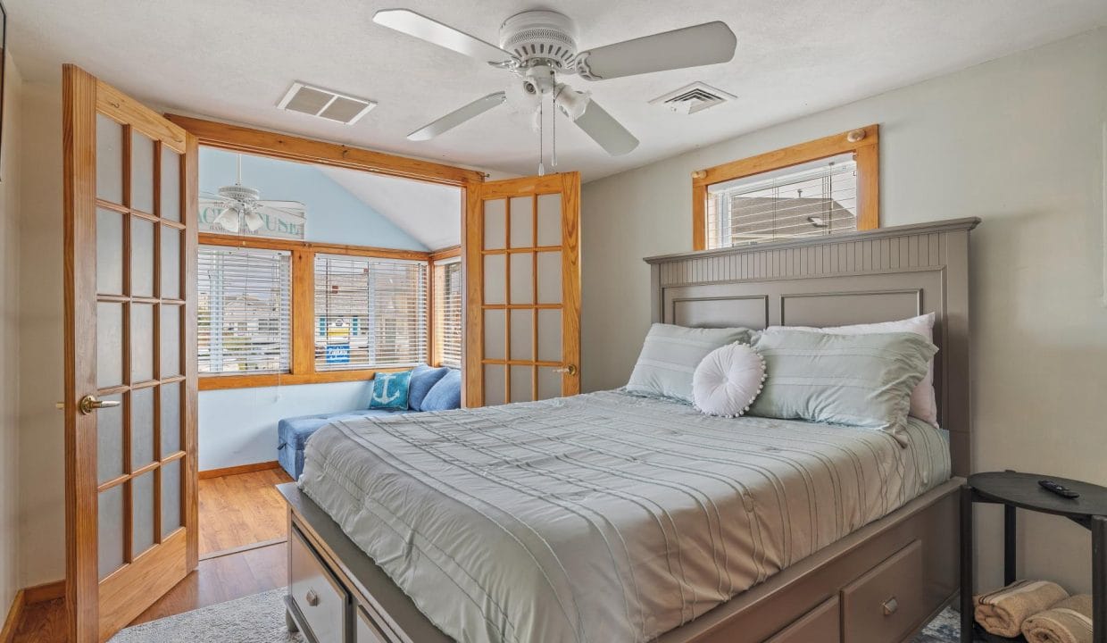 Bedroom with a large bed, light bedding, and a ceiling fan. Wooden doors open to a sunlit nook with a blue chair and wooden flooring.