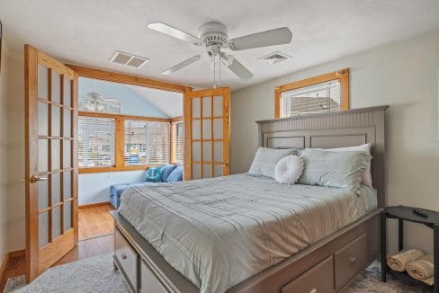 Bedroom with a large bed, light bedding, and a ceiling fan. Wooden doors open to a sunlit nook with a blue chair and wooden flooring.