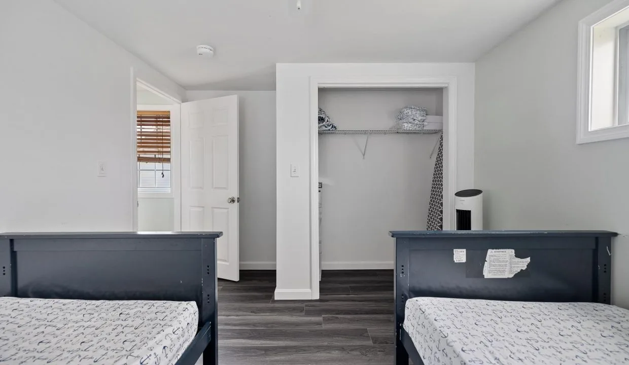 A room with two twin beds, open closet, and a slightly ajar door. Light-colored walls, wooden floor, and a window with blinds.