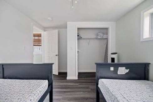 A room with two twin beds, open closet, and a slightly ajar door. Light-colored walls, wooden floor, and a window with blinds.