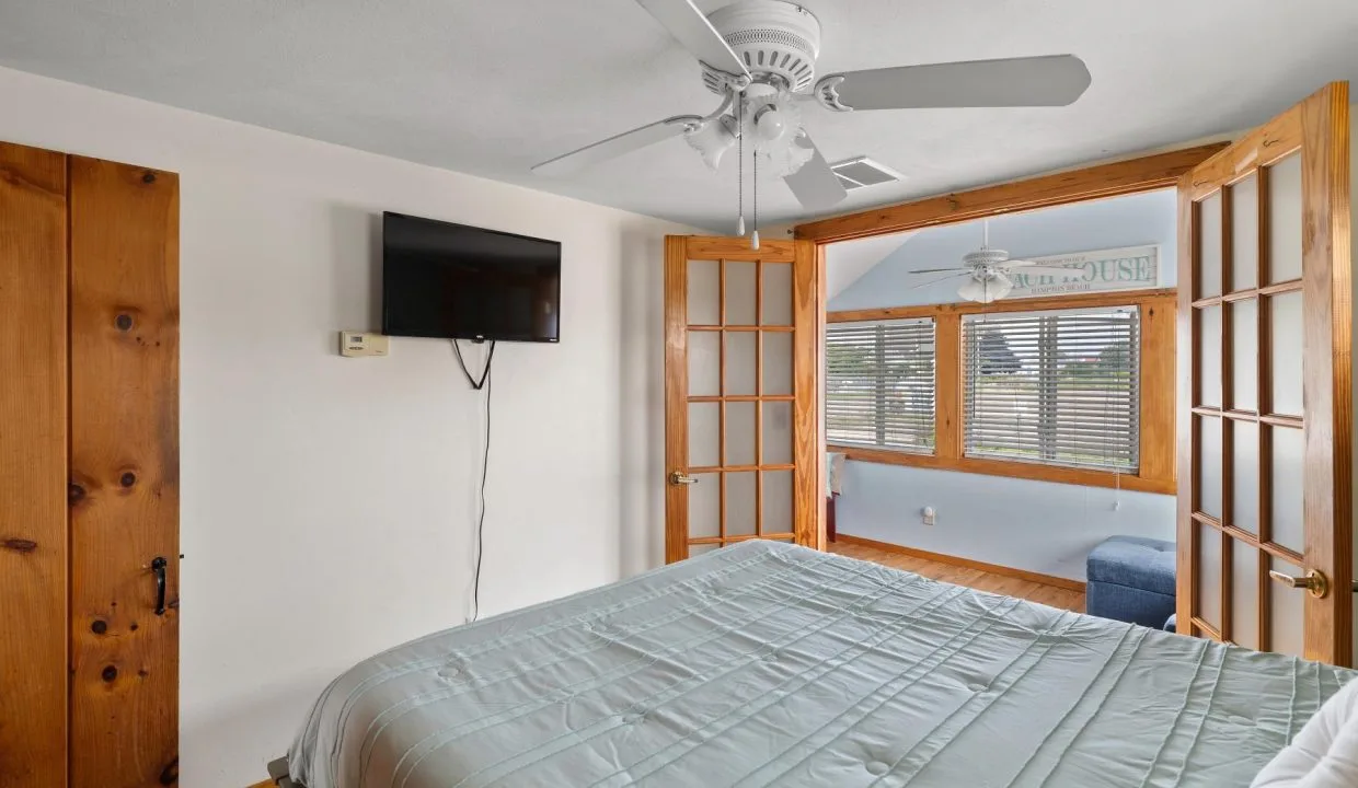 Bedroom with a bed, wall-mounted TV, ceiling fan, and wooden sliding doors leading to a sunlit room with windows and blinds.