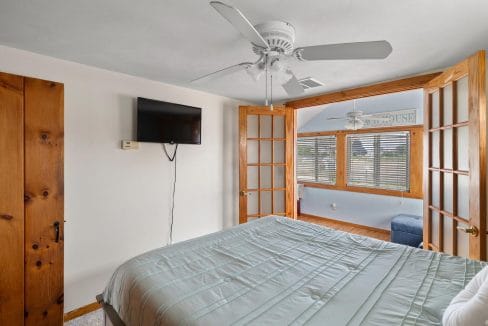 Bedroom with a bed, wall-mounted TV, ceiling fan, and wooden sliding doors leading to a sunlit room with windows and blinds.