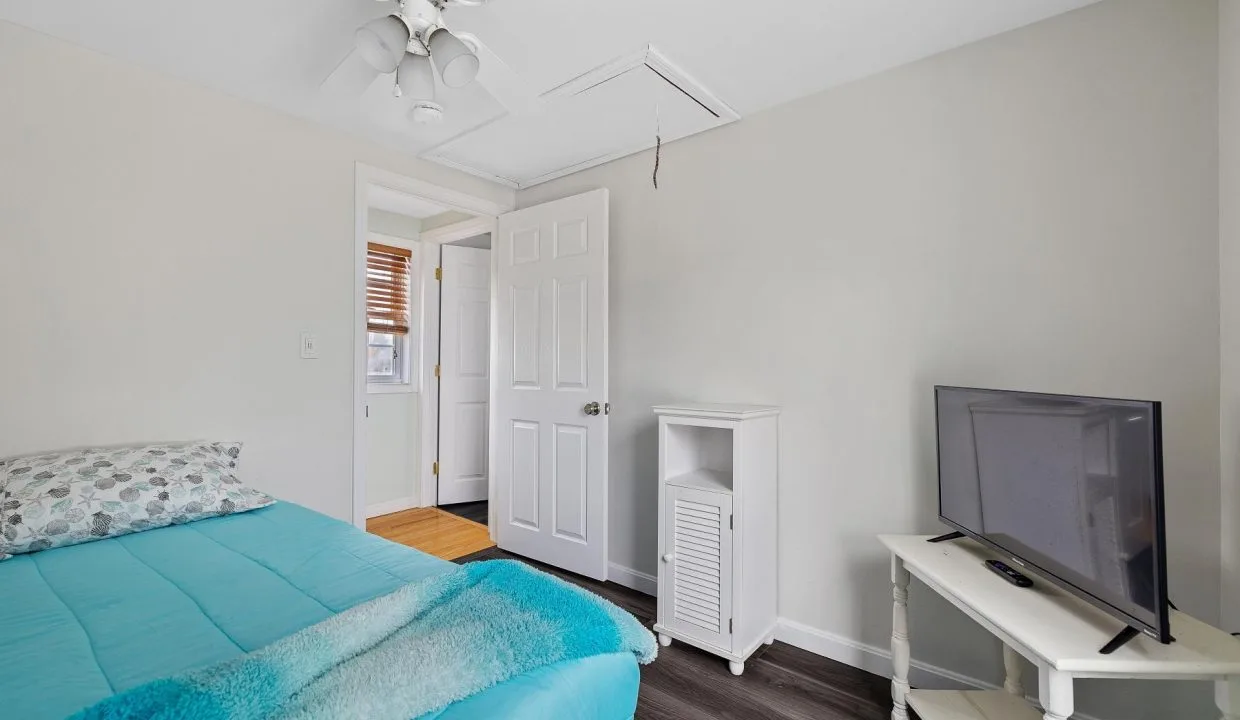 A bedroom with a bed covered in turquoise bedding, a small TV on a white table, and a ceiling fan. There is a door to a hallway and wood flooring is visible.