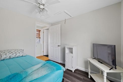 A bedroom with a bed covered in turquoise bedding, a small TV on a white table, and a ceiling fan. There is a door to a hallway and wood flooring is visible.