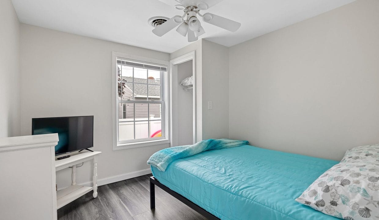 Small bedroom with a single bed covered in turquoise bedding, a TV on a white stand, and a ceiling fan. A window provides natural light, showing a partial view of a neighboring building.