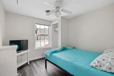 Small bedroom with a single bed covered in turquoise bedding, a TV on a white stand, and a ceiling fan. A window provides natural light, showing a partial view of a neighboring building.