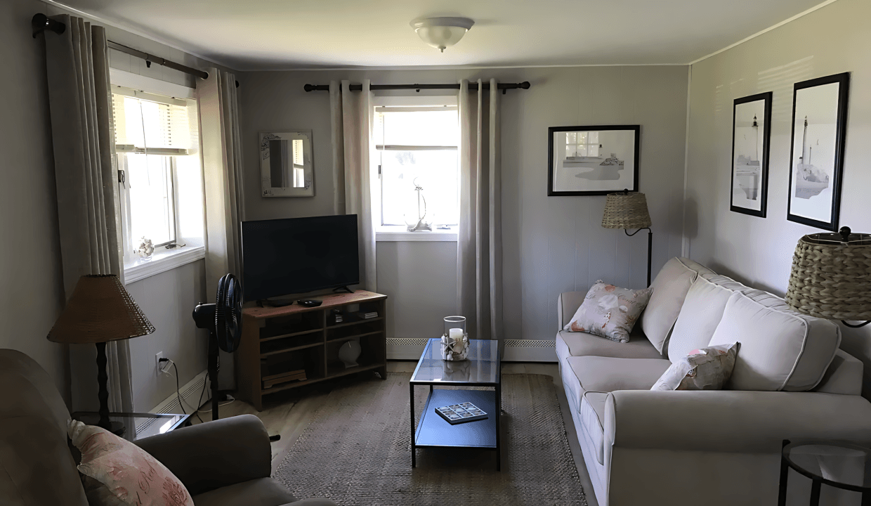 A cozy living room with a beige sofa and armchair, a TV on a wooden stand, a coffee table, and framed artwork on the walls. Natural light filters through curtained windows.