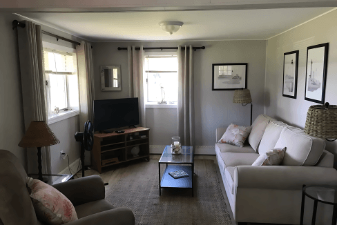 A cozy living room with a beige sofa and armchair, a TV on a wooden stand, a coffee table, and framed artwork on the walls. Natural light filters through curtained windows.