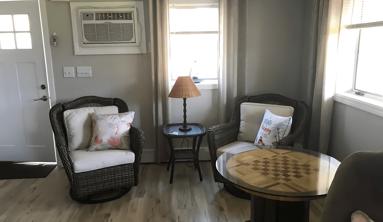 Cozy living room with two wicker chairs, a side table with a lamp, and a round table featuring a chessboard pattern. A window and an air conditioner are on the back wall.