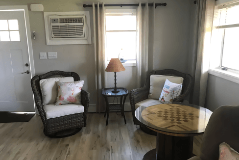 Cozy living room with two wicker chairs, a side table with a lamp, and a round table featuring a chessboard pattern. A window and an air conditioner are on the back wall.