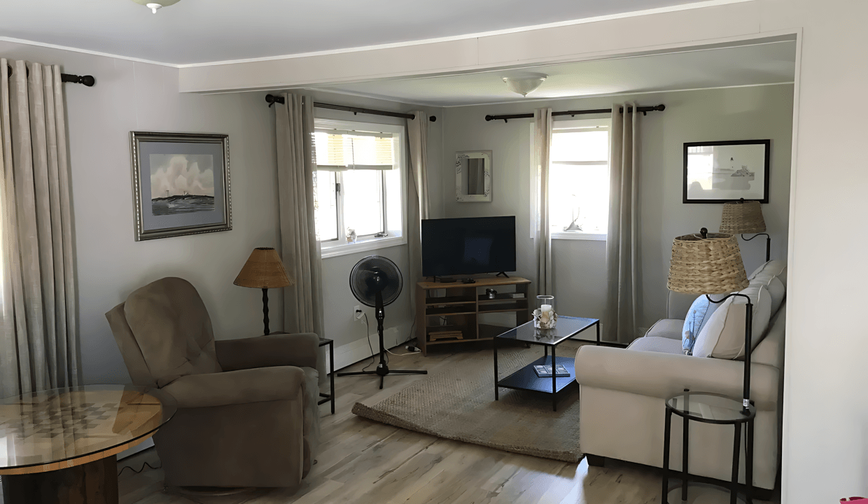 Living room with beige walls, two armchairs, a TV on a wooden stand, a circular table, and a ceiling light. Curtains cover windows, and a fan is near a chair.