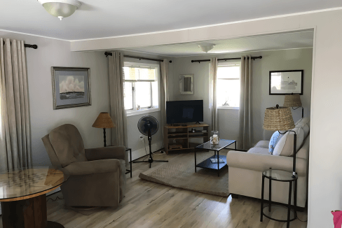 Living room with beige walls, two armchairs, a TV on a wooden stand, a circular table, and a ceiling light. Curtains cover windows, and a fan is near a chair.