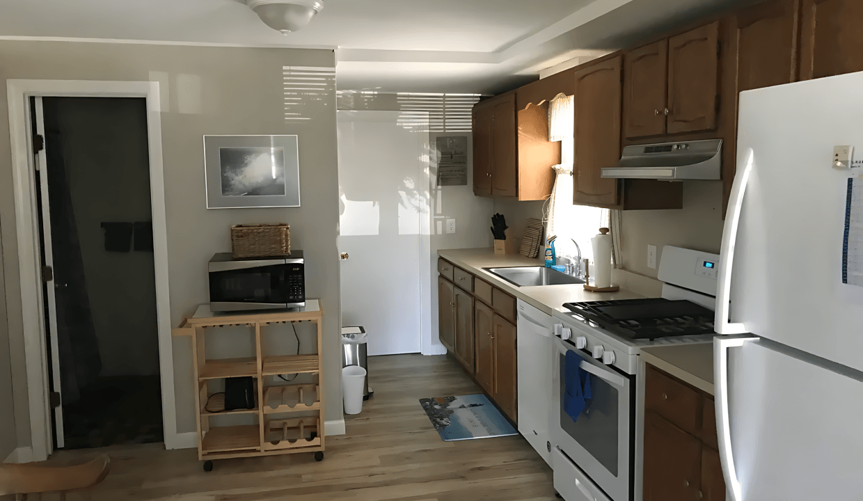 A kitchen with wooden cabinets, a white refrigerator, stove, microwave, and a rolling cart. There's a doorway to another room and natural light from a window.