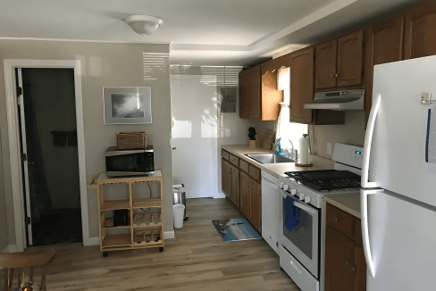 A kitchen with wooden cabinets, a white refrigerator, stove, microwave, and a rolling cart. There's a doorway to another room and natural light from a window.