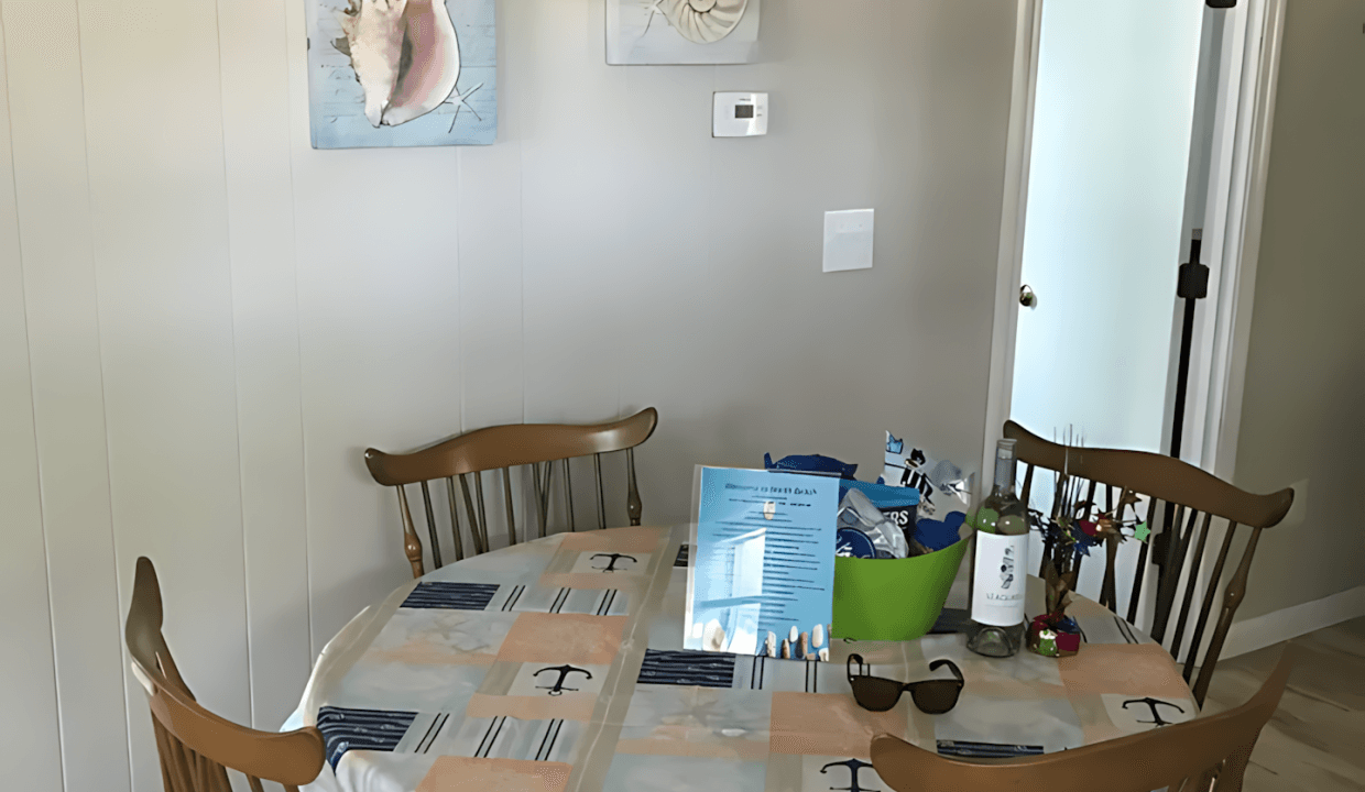 A dining area with a wooden table and four chairs, covered by a patterned tablecloth. A hanging light fixture is above the table. Two shell-themed pictures adorn the beige wall.