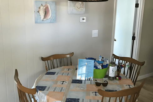 A dining area with a wooden table and four chairs, covered by a patterned tablecloth. A hanging light fixture is above the table. Two shell-themed pictures adorn the beige wall.