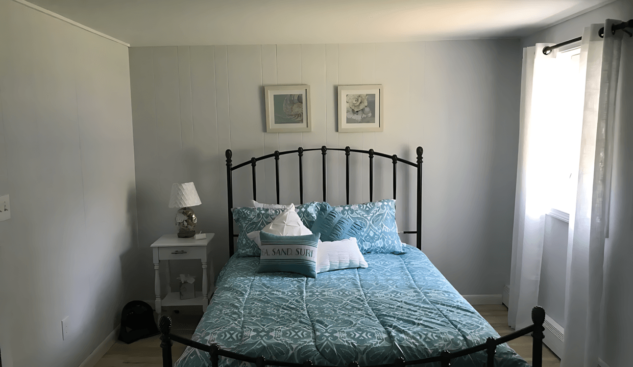 Bedroom with a metal bed frame, blue patterned bedding, and two pillows. A small white nightstand with a lamp and two paintings are on the wall. Light streams through a window with curtains.