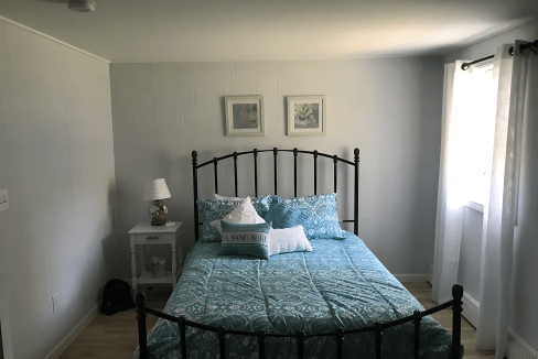 Bedroom with a metal bed frame, blue patterned bedding, and two pillows. A small white nightstand with a lamp and two paintings are on the wall. Light streams through a window with curtains.