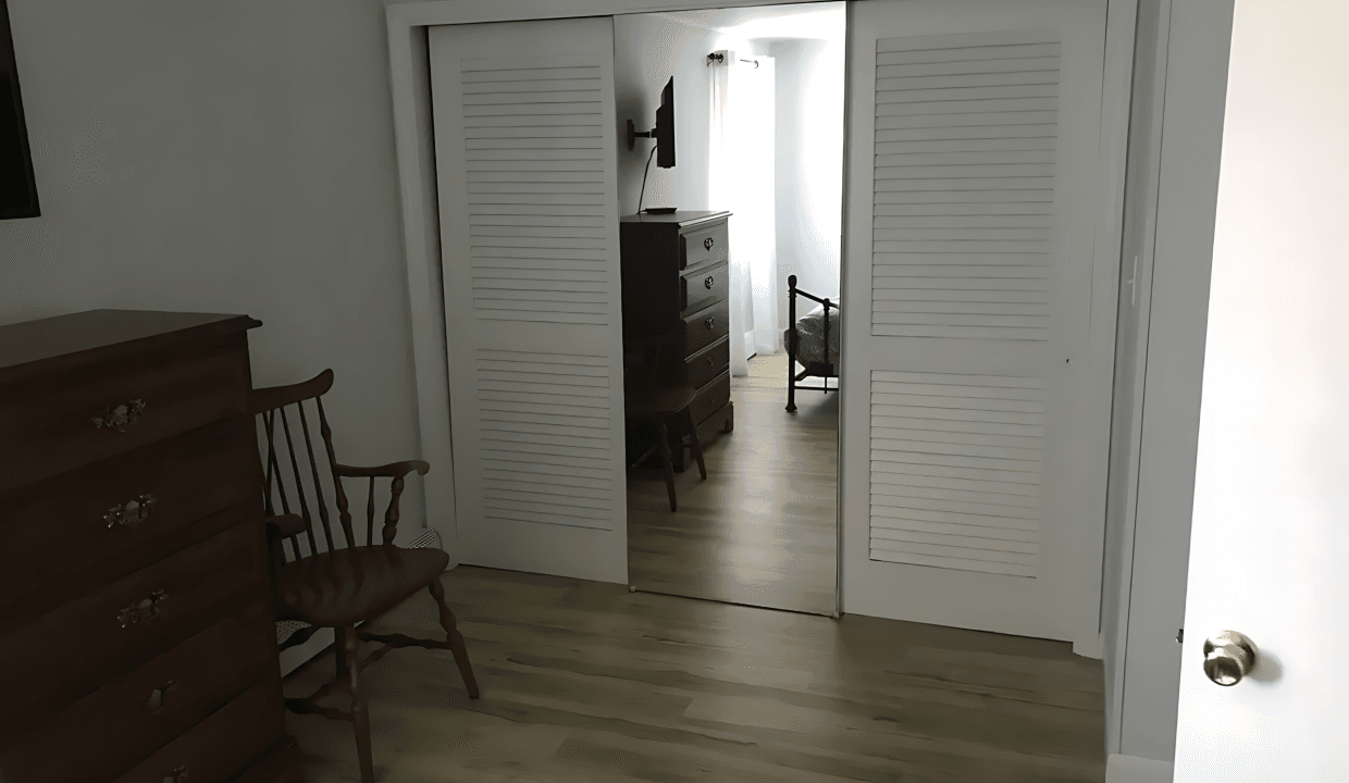 Room with wooden floor, dresser, and chair. Sliding closet doors partially open, revealing another room with a chest of drawers and natural light.