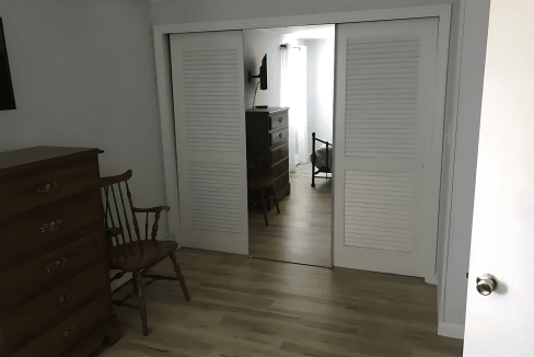 Room with wooden floor, dresser, and chair. Sliding closet doors partially open, revealing another room with a chest of drawers and natural light.