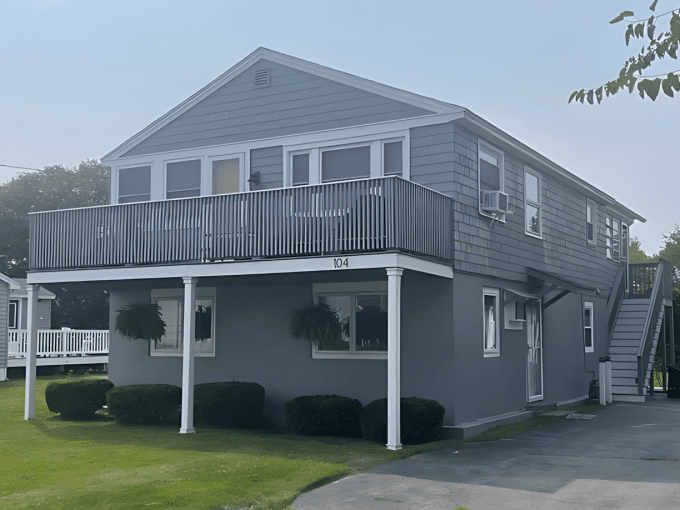 Two-story gray house with an upper-level balcony, front lawn, and external staircase. The number "104" is displayed on the front.